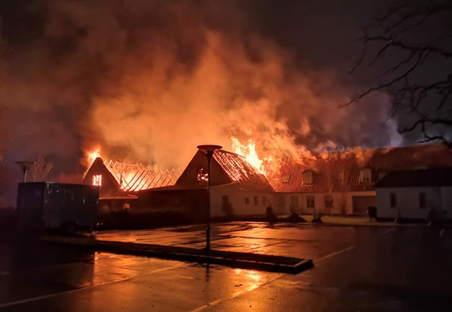 En voldsom brand i en stor bygning om natten, hvor flammer og røg stiger op fra taget. Gårdspladsen foran bygningen er oplyst af ildens skær og reflekterer på den våde asfalt.