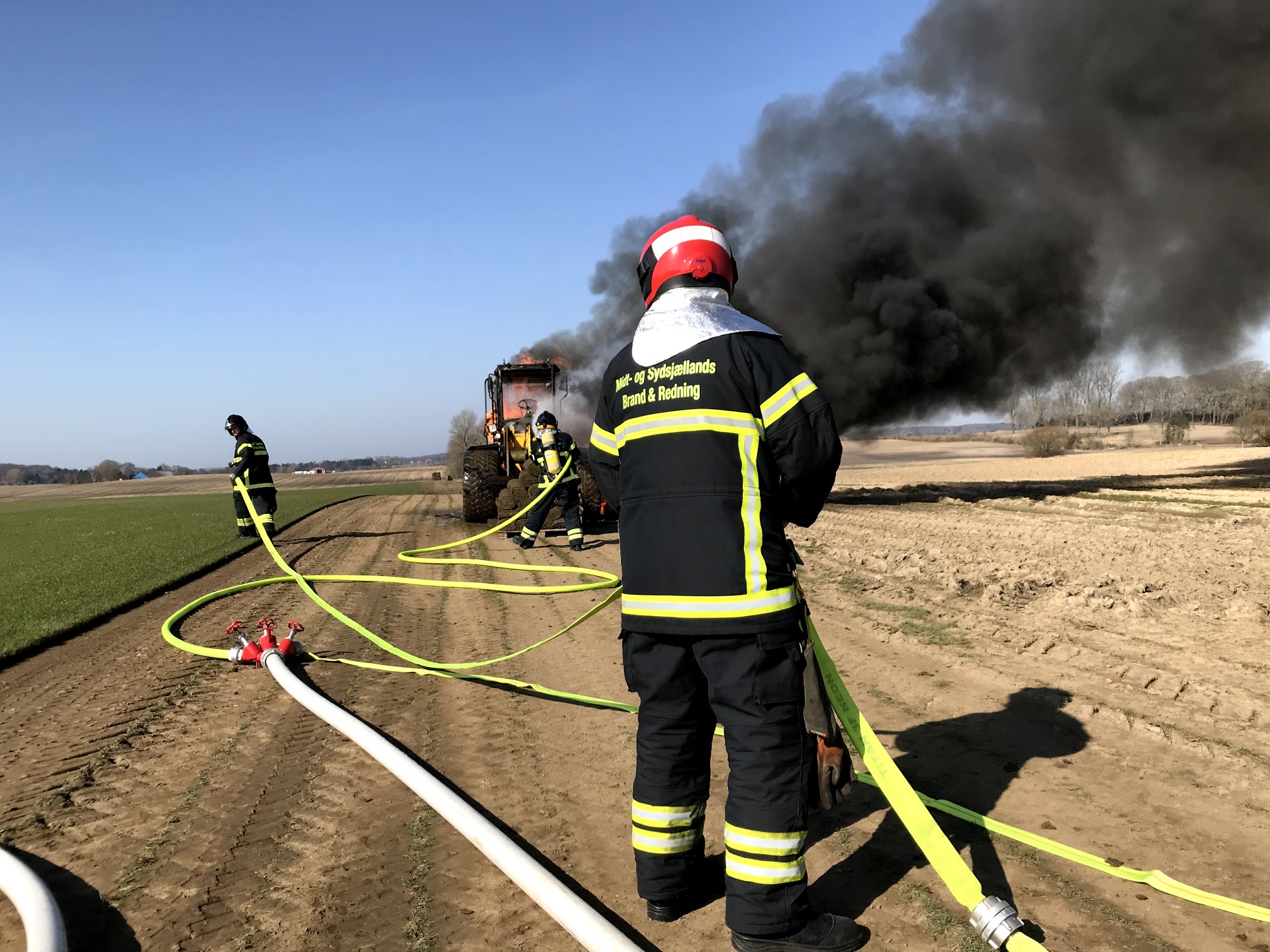 Brandfolk fra Midt- og Sydsjællands Brand & Redning slukker en brand i en traktor på en mark. Sort røg stiger op fra traktoren, mens brandslanger ligger udstrakt på marken.