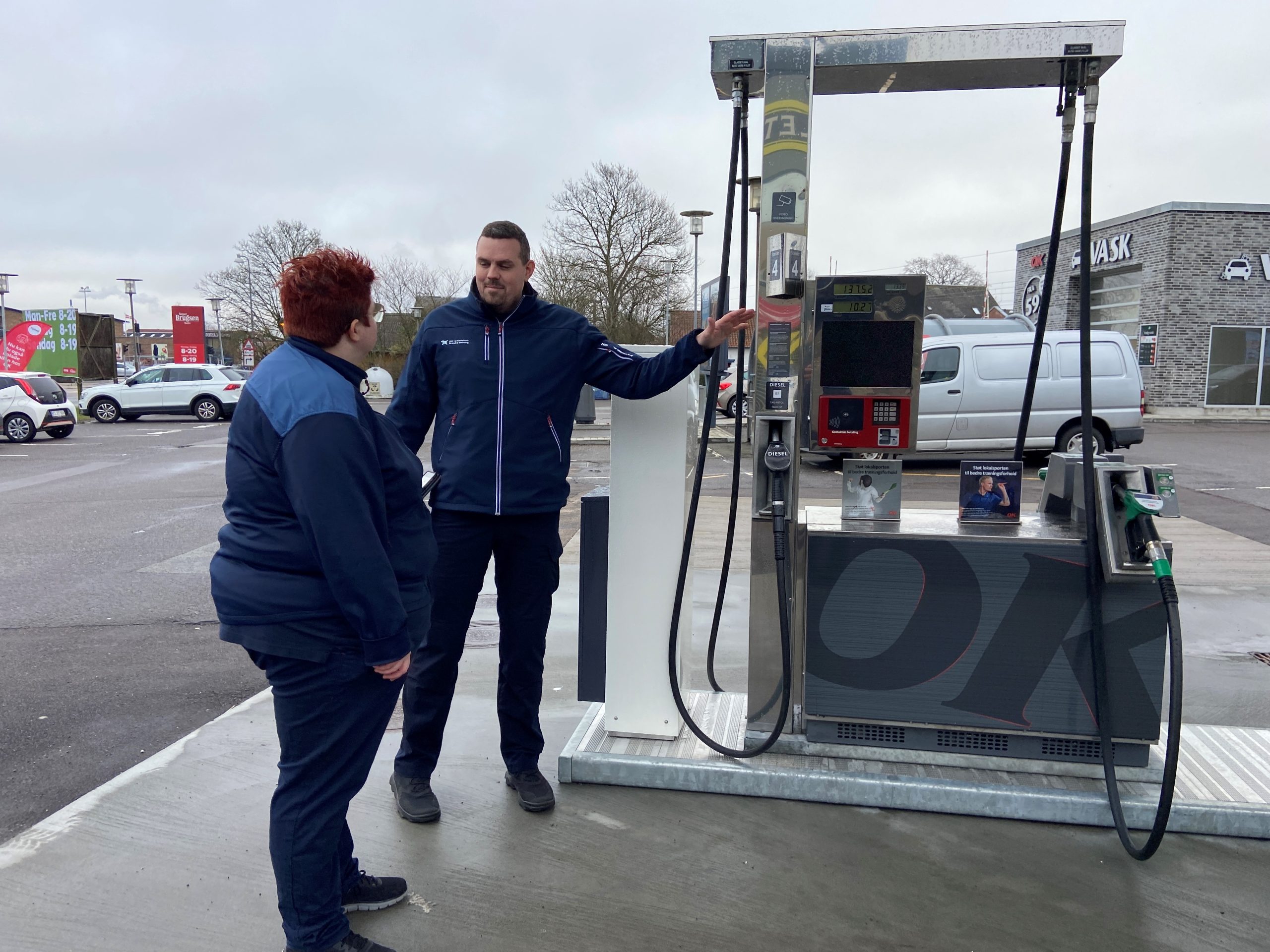 En brandinspektør udfører et brandsyn ved en OK-tankstation og er i samtale med en medarbejder. Han gestikulerer mod brændstofstanderen, mens medarbejderen lytter opmærksomt. Bag dem ses parkerede biler, en vaskehal og et butiksskilt med åbningstider. Den grå, overskyede himmel danner en neutral baggrund for inspektionen.