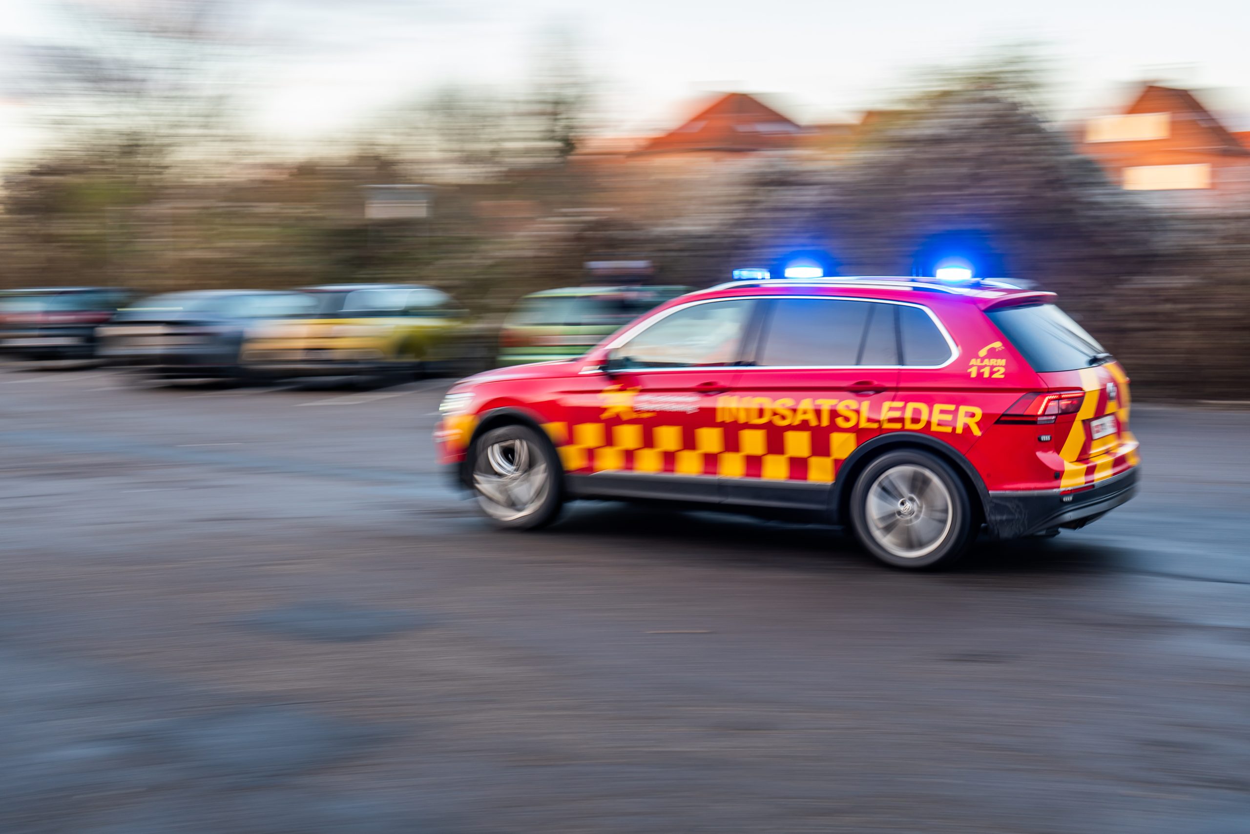 En indsatslederbil i rødt og gult med blå blink kører hurtigt gennem et boligområde, med bevægelsesuskarphed i baggrunden.