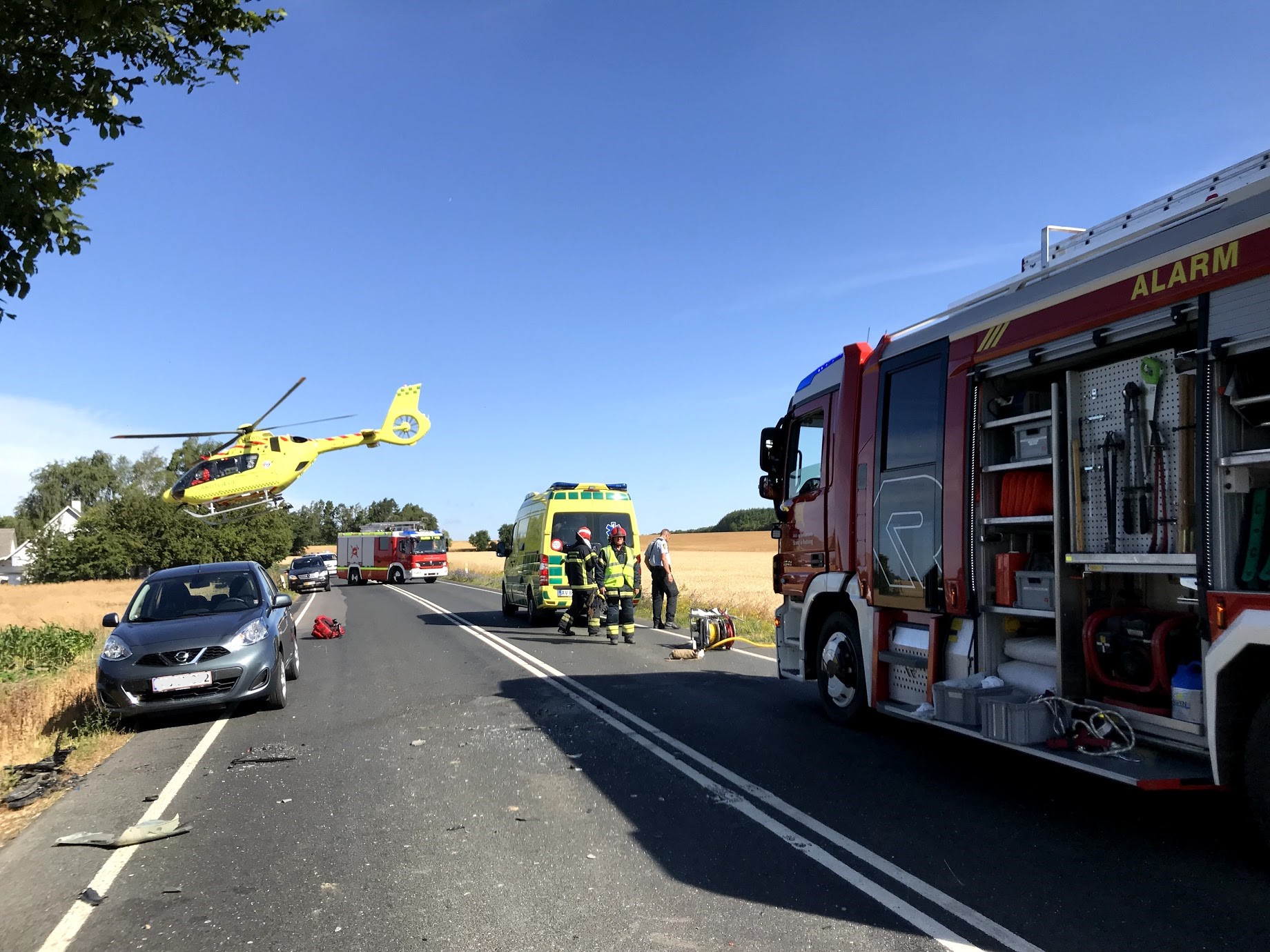 Redningsoperation på en landevej med en helikopter, brandbil, ambulance og redningsfolk, der arbejder ved en trafikulykke. En bil er parkeret i vejkanten, og marker kan ses i baggrunden.