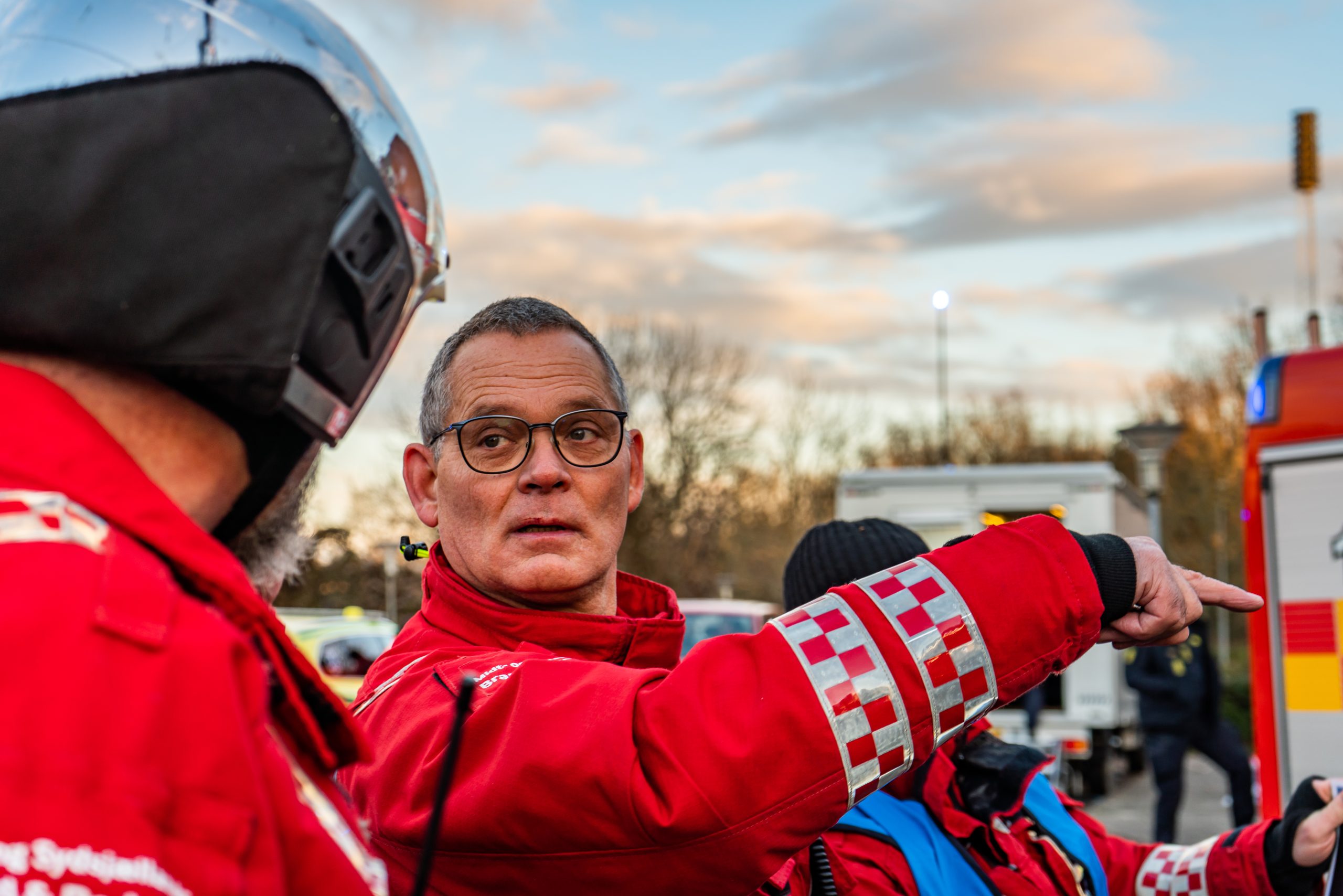 Redningsleder i rød uniform peger mod et område under en redningsaktion, mens han kommunikerer med en kollega iført hjelm. Baggrunden viser træer og køretøjer under en himmel med skyer.