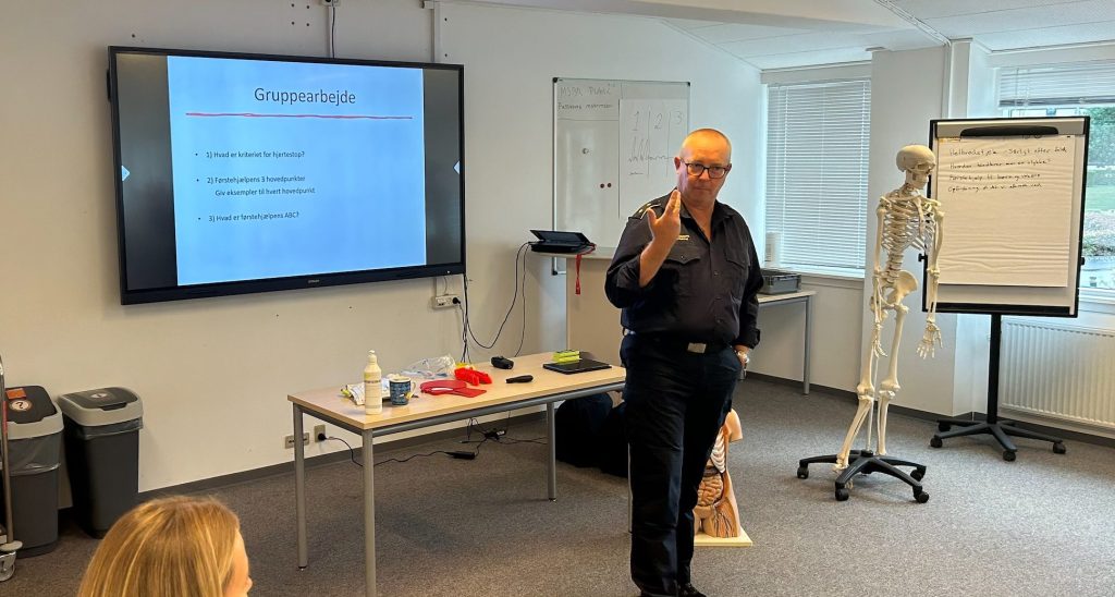 Førstehjælpsinstruktør iført uniform står foran powerpoint præsentation og underviser i førstehjælp. Der ses et anatomisk plastik skelet, forskellige førstehjælpsremedier og andre almindelige undervisningslokale-genstande.