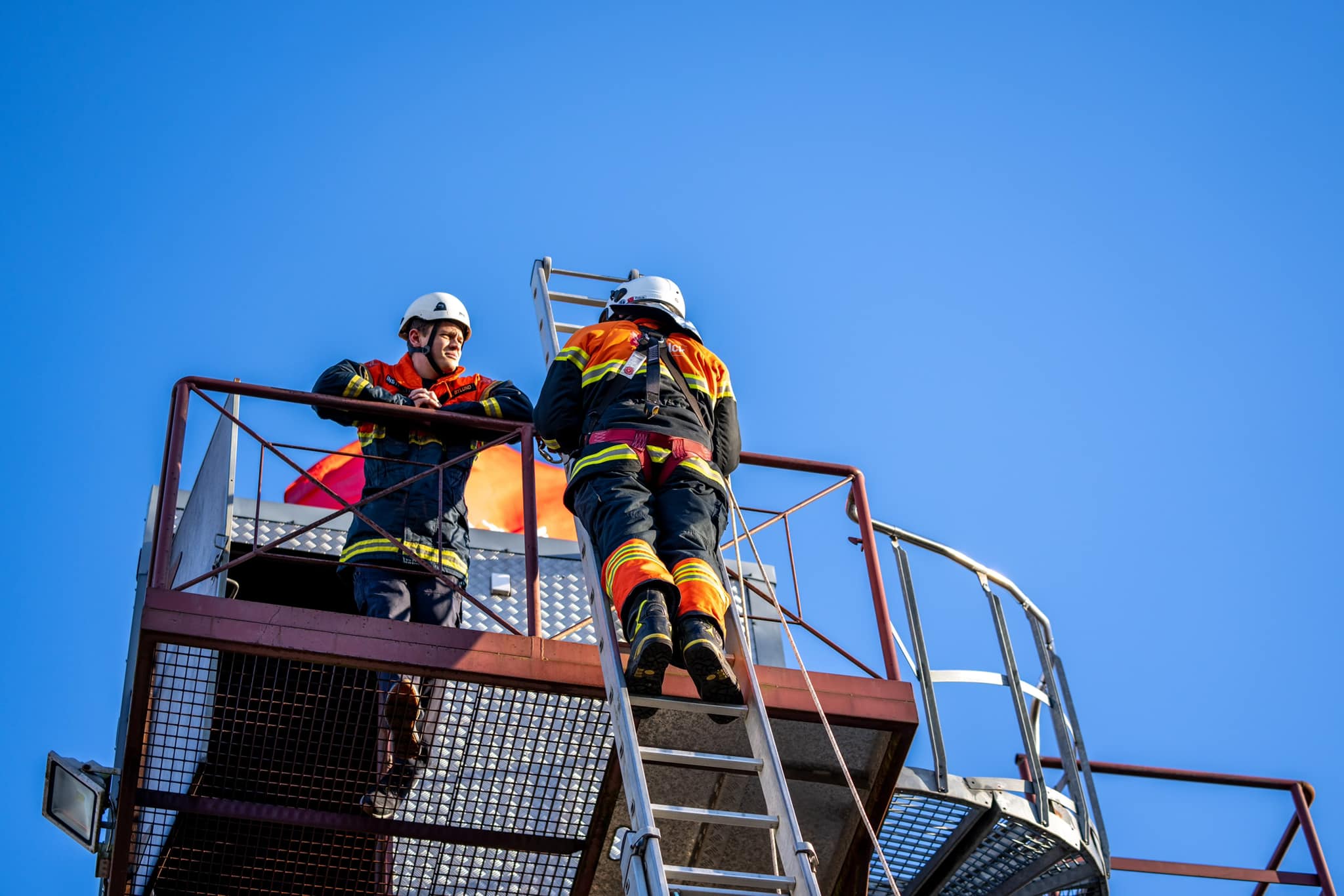 To brandmmænd, den ene står højt oppe på en stige med en faldsikringssele på imens han snakker med den anden brandmand som står på en form for plateau.