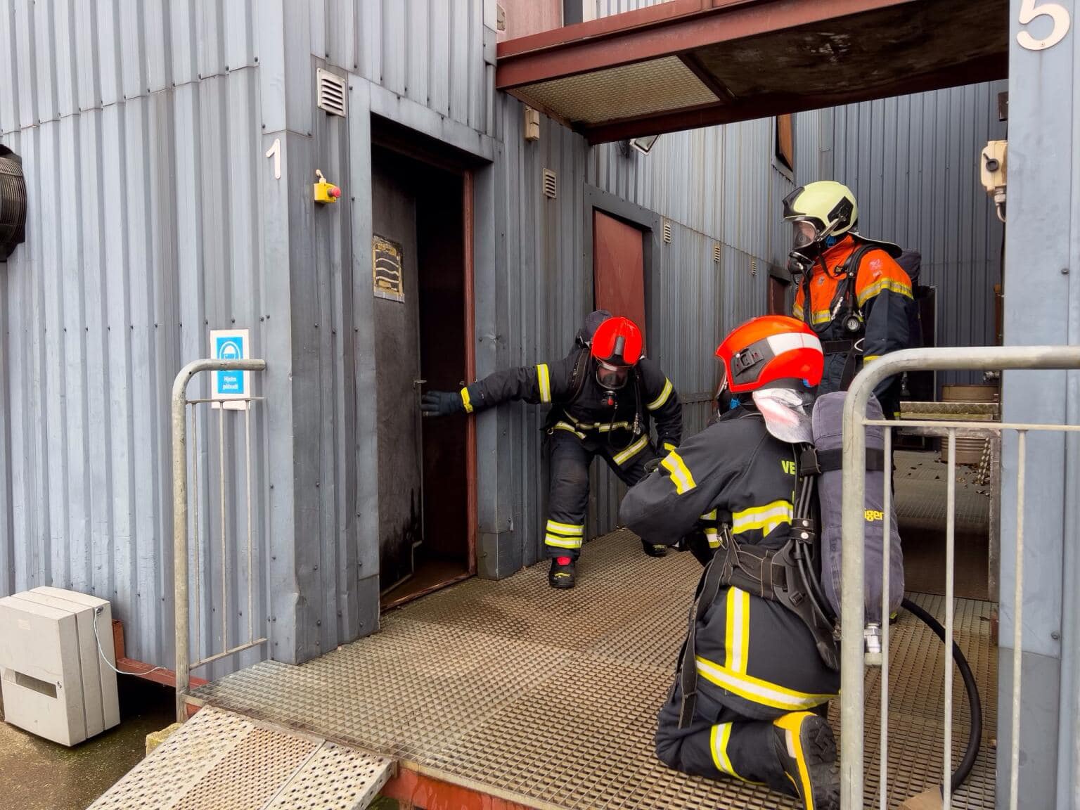 Tre brandfolk iført røgdykkerudstyr og hjelme deltager i en øvelse på en brandskole. En af dem åbner forsigtigt en dør til en metalbygning, mens en anden knæler med en brandslange klar. En tredje brandmand, iført en orange jakke, overvåger situationen bag dem. Øvelsen foregår på en hævet metalplatform med gelænder, og bygningen har synlige mærkater og instruktioner.