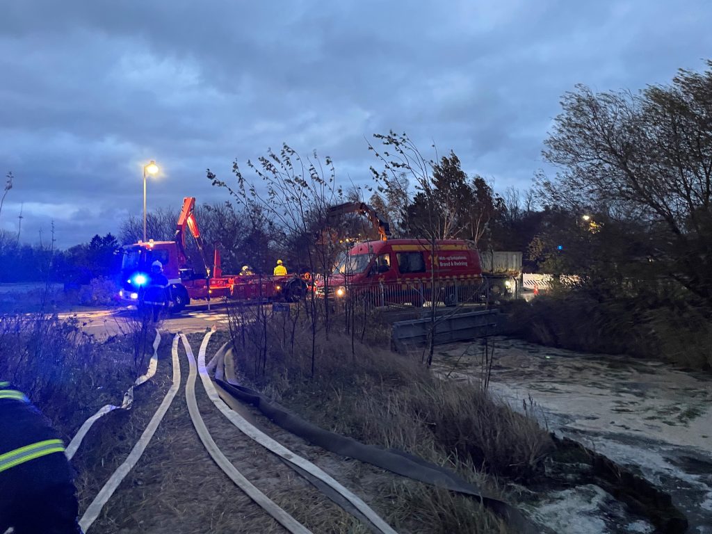 Brand- og redningsfolk arbejder ved skumfyldt vandløb i skumringen. Brandslanger er lagt ud på jorden, og en rød brandredningsvogn med kran er til stede. Blå blink lyser op i mørket, og redningspersonale bærer refleksveste og hjelme.