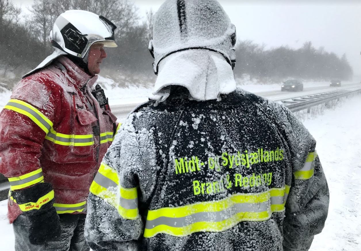 To brandfolk fra Midt- og Sydsjællands Brand & Redning står i snevejr ved en motorvej. Deres beskyttelsesdragter og hjelme er dækket af is og sne. Den ene brandmand har ryggen til kameraet, og teksten 'Midt- og Sydsjællands Brand & Redning' er synlig på jakken. Den anden brandmand står i profil, iført en rød jakke med reflekser. I baggrunden kan man se en sneklædt vej med biler, der passerer forbi.