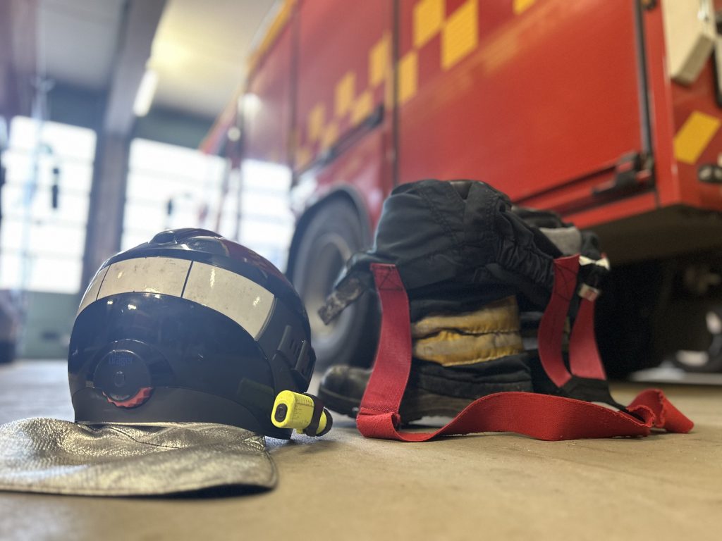 Brandhjelm og beskyttelsesudstyr ligger klar på gulvet foran en rød brandbil i garage.