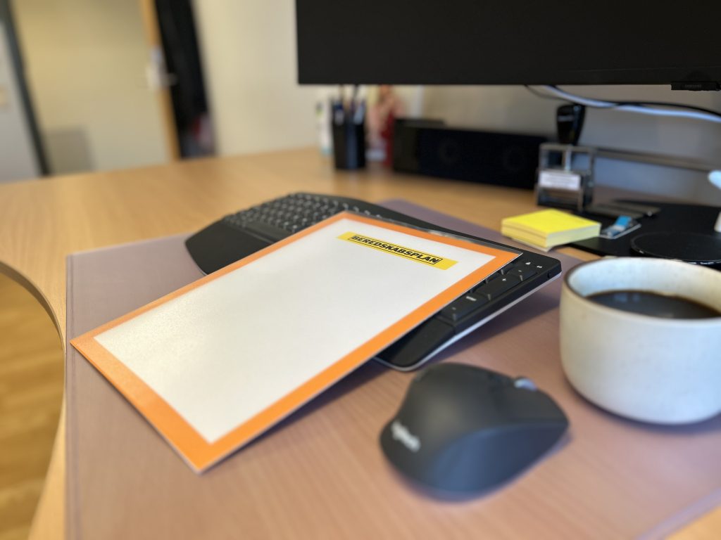 Dokument med titlen 'Beredskabsplan' ligger på et skrivebord ved siden af en computer, mus og kaffekop.